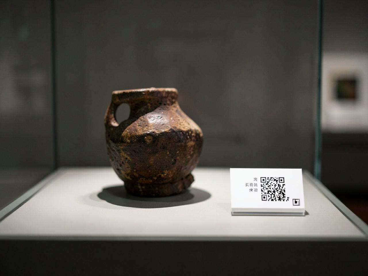 A single antique vessel lit in a museum case beside its existing white label card carrying a small code